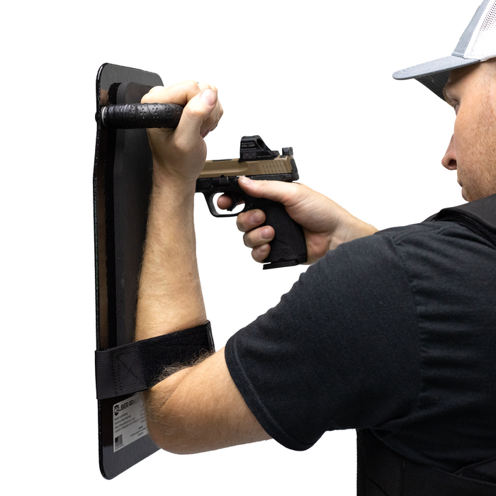 Person holding up a black 8" x 17" Caliber Armor Ballistic Shield with handle padded armrest & forearm strap while pointing a pistol 6